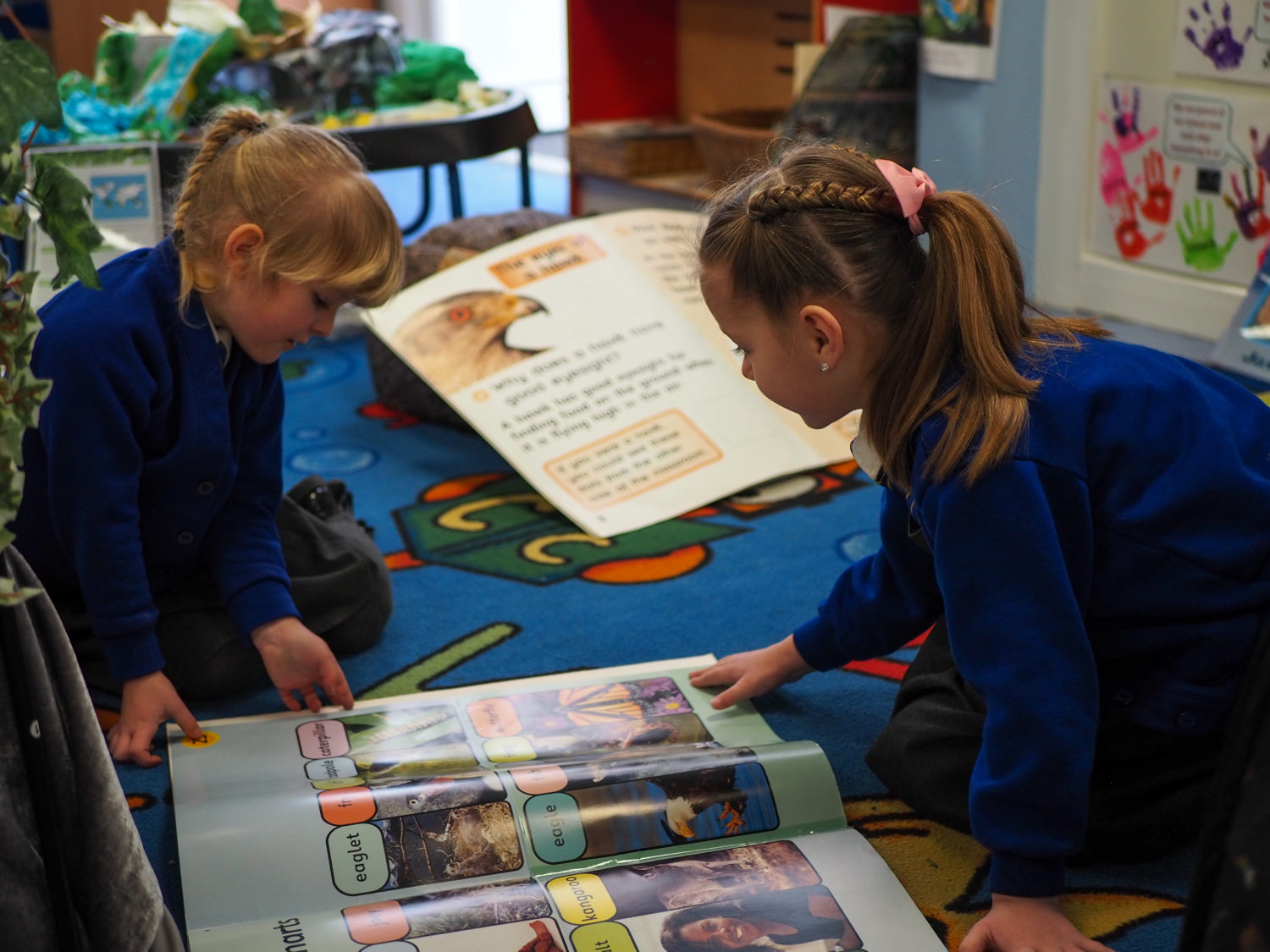 Two young girls are shown sitting on the carpet in a classroom, looking at some picture storybooks together.