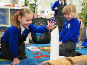 Two young girls are pictured sat together on a carpeted area in a classroom, reading some picture storybooks together.