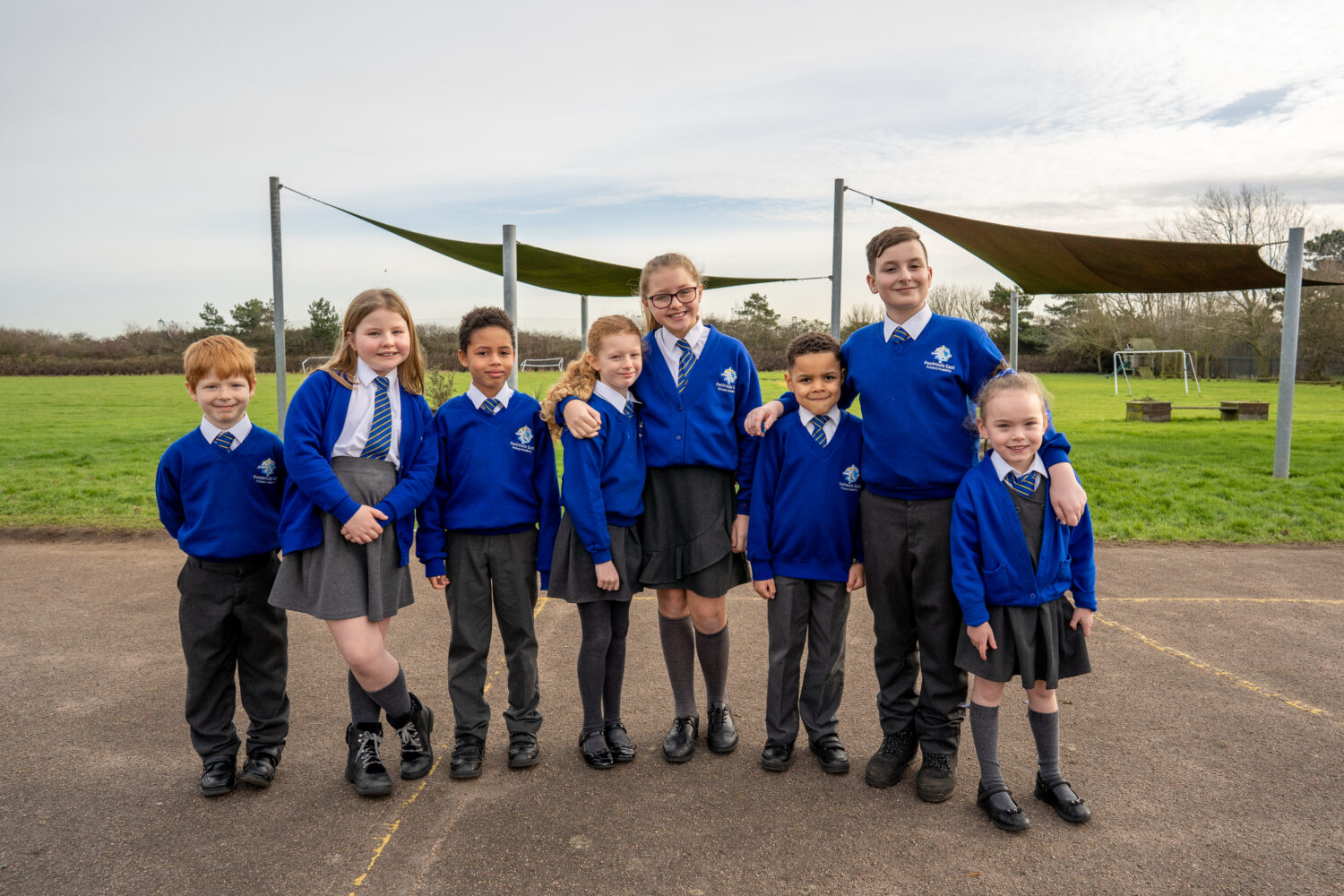 Students from various year groups are pictured huddled together for a group photo, outdoors on the academy grounds, smiling brightly.